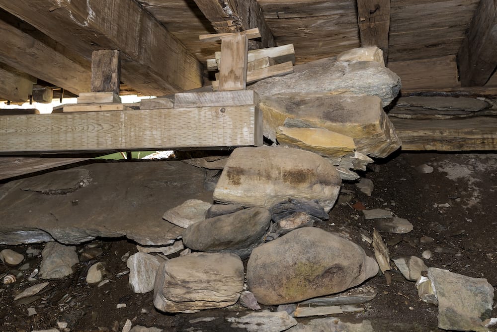 A pile of rocks, blocks of wood, and a wooden beam hold up a floor in an unfinished basement. Floor is dirt and rock. Outside light can be seen through hole. (A pile of rocks, blocks of wood, and a wooden beam hold up a floor in an unfinished basement